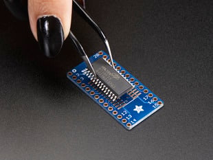 A pair of tweezers holding a microchip hovers over the SMT Breakout PCB for SOIC-28 or TSSOP-28.