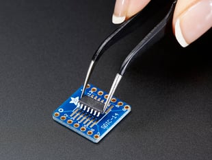 A pair of tweezers holding a microchip hovers over the SMT Breakout PCB for SOIC-14 or TSSOP-14.