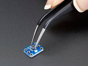 A pair of tweezers holding a microchip hovers over a small rectangular Breakout PCB.