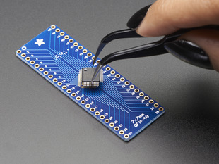 A pair of tweezers holding a microchip hovers over the SMT Breakout PCB for 48-QFN or 48-TQFP placement on a long blue rectangular breakout board.