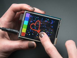 Brown polished fingers holding a 2.8" TFT Touch Shield for Arduino with Resistive Touch Screen with one hand and a finger from the other hand drawing a heart. 