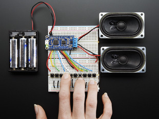 A long blue rectangular audio FX sound board assembled onto two half-size breadboards. The board is wired up to a 3 x AA battery pack and two speakers. A white hand presses a row of small tactile buttons wired up to the audio board.