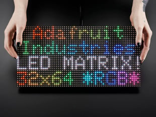 Two white hands hold out an assembled and powered on 64x32 RGB LED Matrix Panel - 6mm pitch. The matrix displays "Adafruit Industries LED MATRIX! 32x64 *RGB*"