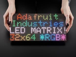 Two white hands hold out an assembled and powered on 64x32 RGB LED Matrix Panel - 5mm pitch. The matrix displays "Adafruit Industries LED MATRIX! 32x64 *RGB*"