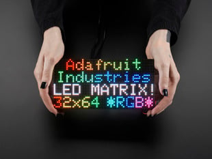 Two white hands hold out an assembled and powered on 64x32 RGB LED Matrix Panel - 3mm pitch. The matrix displays "Adafruit Industries LED MATRIX! 32x64 *RGB*"