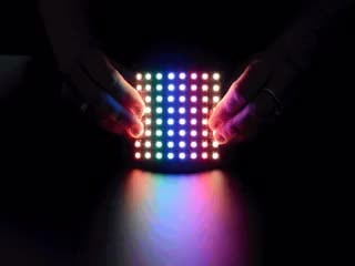 Two white hands holding a rainbow pulsing Flexible 8x8 NeoPixel RGB LED Matrix.