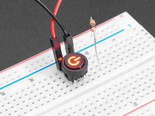 Close-up of a mini red illuminated push-button with a red power symbol installed in a half-size breadboard. Also are on the breadboard are two jumper wires and a resistor.