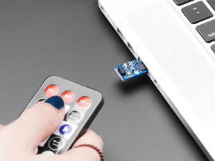 A Python Programmable InfraRed USB Adapter plugged into a macbook and a blue-manicured hand holding a black rectangular remote. 
