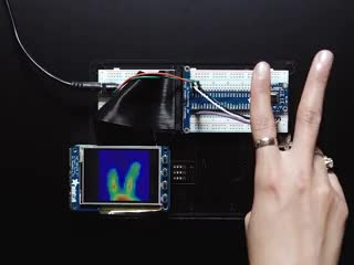 Video of a hand making bunny ears with their fingers over the AMG8833 IR thermal sensor. A thermal image readout displays on a TFT screen wired into a breadboard.