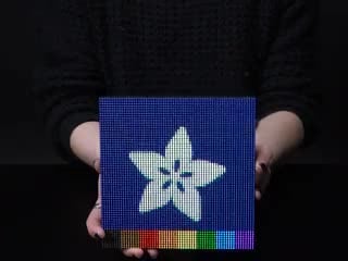 Video of two hands flipping over a powered on 64x64 RGB LED Matrix. The matrix display has a falling colored sand effect. 