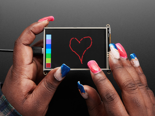 Overhead shot of a Black woman's hands with a blue and pinkish-red manicure drawing a heart on a touchscreen breakout.