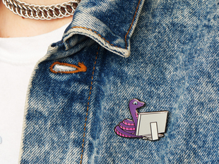 Lifestyle image of a denim jacket on top of a white shirt showing off an enamel pin resembling a purple-and-pink python using a desktop computer.
