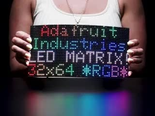 Video of woman in a white sleeveless t-shirt holding and slightly bending a black flexible 64x32 RGB LED Matrix. The LED matrix is lit up with rainbow LEDs with the text: Adafruit Industries LED Matrix 32x64 *RGB*.