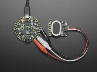 Top view of a black speaker breakout board connected to a round microcontroller via alligator clips. A music note animation emits from the speaker.