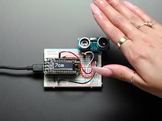Video of a woman's hand rising above a blue ultrasonic sonar distance sensor plugged into a half-size breadboard. An OLED screen displays the rising distance between the woman's hand and the sensor.