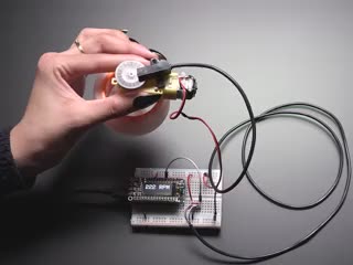 Hand holding motor with spinning wheel. T-slot sensor is attached over an encoder disk and is showing the RPM on an OLED display