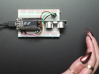 Video of a woman's hand moving toward ultrasonic sonar distance sensor plugged into a half-size breadboard. An OLED screen displays the distance between the woman's hand and the sensor.