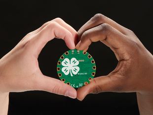 A white hand and a Black hand meet at the center of the frame to create a heart symbol. They hold up a round green dev board with the 4-H logo in white.