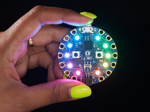 shot of a Black woman's neon-green manicured hand holding up a Circuit Playground Bluefruit glowing rainbow LEDs.