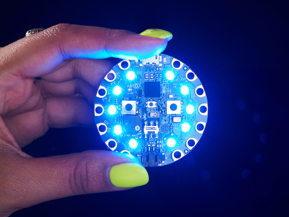 shot of a Black woman's neon-green manicured hand holding up a Circuit Playground Bluefruit glowing blue LEDs.
