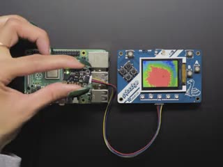White hand holding and moving around a MLX90640 24x32 IR Thermal Camera Breakout over a Raspberry Pi. The pybadge showing a checker board image of what the camera is seeing. 