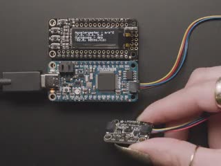 Video of a white hand moving a sensor around that connected to an OLED and a blue rectangular board. 