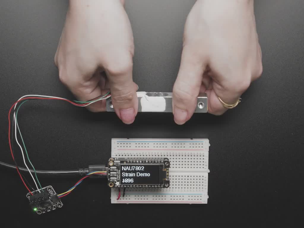 Video of a pair of white hand's trying to bend a strain gauge. The gauge is connected to a breakout board which is also connected to an OLED display on a half-size breadboard. The numbers on the OLED display show high digit numbers.