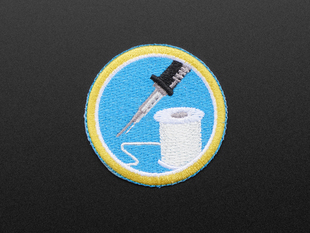 Circular embroidered badge with image of soldering iron and solder spool on blue background with yellow edge. 