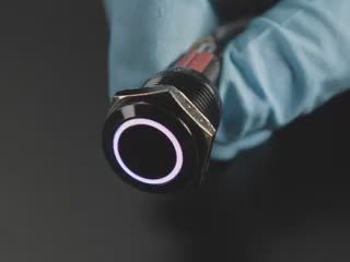 Angled video of 16mm rugged black metal pushbutton with LED ring glowing rainbow colors. A person with gloved hands holds the button and pushes the switch on and off.