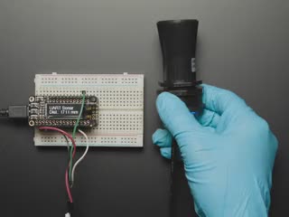 A blue-gloved hand articulates a black ultrasonic sensor horn connected to an OLED display on a half-size breadboard. The OLED displays distance data.
