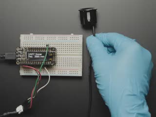 A blue-gloved hand articulates a small black ultrasonic sensor horn connected to an OLED display on a half-size breadboard. The OLED displays distance data.