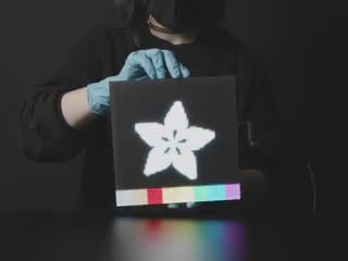 Video of a woman in black slowly rotating a powered-on 64x64 RGB LED Matrix. The matrix displays the Adafruit star flower logo with rainbow sand falling animation.
