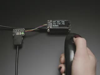 Hand holding a Wiichuck controller pressing buttons. The controller is connected to the breakout wired to a Feather with OLED showing the streaming controller data