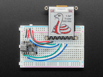 Top view of a 1.54" tri-color eInk display assembled on a breadboard with jumper wires and a QT Py. Friendly snake in white-and-red, Blinka, on the display.