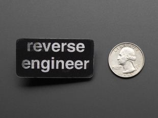 straight down view of black rectangular badge with the words REVERSE ENGINEER in white, next to a quarter for scale. 