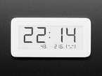 Top view of white rectangular digital clock with electronic ink display. The display reads the time, 22:14, along with 43% humidity, 21.6ºC with a smiley emoticon.