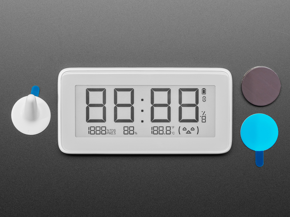 Top view of white rectangular digital clock with electronic ink display next to a circular magnet and two mounting stickers.