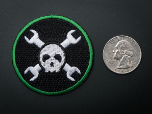 Circular embroidered badge with white skull over two crossed wrenches on black background with green trim. Next to quarter for scale. 