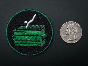 Circular embroidered badge with green dumpster and a white stick figure mid-dive, on black background with green trim. Shown next to a quarter for scale. 