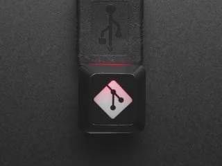 Top view video of black GIT logo keycap connected via USB cable. Various colors emit through the logo.