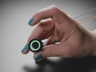 Video of a cyan blue-manicured hand pressing a latching push-button. The LED ring around the push-button glows rainbow colors.