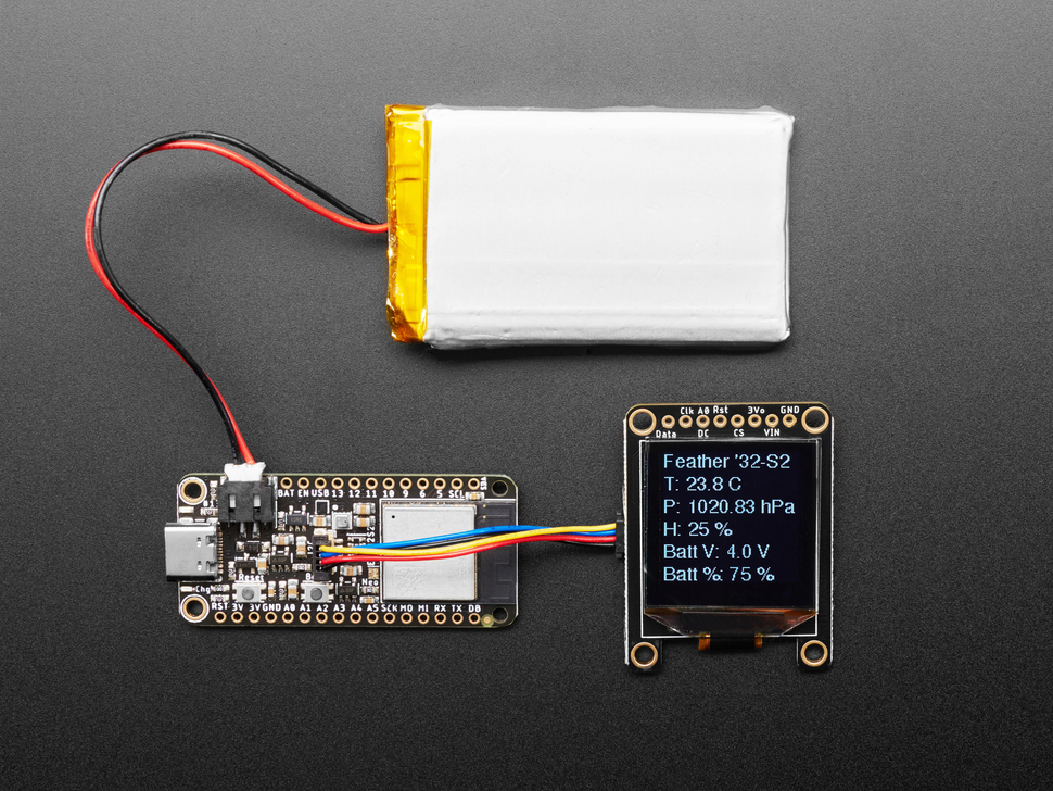 Top view of rectangular microcontroller connected to a LiPo battery and a monochrome OLED display. The OLED screen displays battery information.