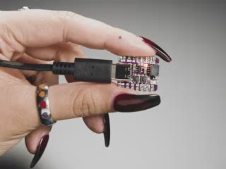 Video of a white hand with long dark red nails holding a small square dev board. The onboard LED glows rainbow colors.