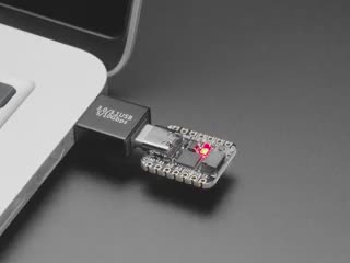 Angled video of a USB adapter plugged into a laptop. A square-shaped microcontroller is plugged into the other end of the adapter. An RGB LED on the microcontroller emits a rainbow spectrum of colors.