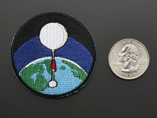 Circular badge with white high altitude balloon over a green and blue earth on a black background, with black trim. Shown next to a quarter for scale. 