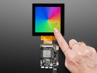 Overhead shot of a 3.4" square TFT screen connected to a microcontroller. The screen displays a colorful rainbow gradient. A white hand draws a heart on the screen.