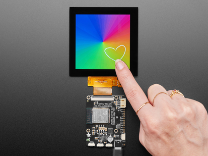 Overhead shot of a 3.4" square TFT screen connected to a microcontroller. The screen displays a colorful rainbow gradient. A white hand draws a heart on the screen.