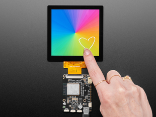 Overhead shot of a 4" square TFT screen connected to a microcontroller. The screen displays a colorful rainbow gradient. A white hand draws a heart on the screen.