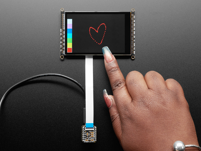 Overhead shot of a Black woman's green-manicured hand touching a 3.5" TFT display breakout connected to a small, square-shaped microcontroller. The TFT screen displays a drawn red heart.