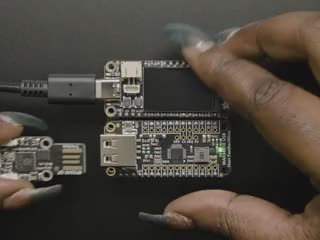 Video of a Black woman's green-manicured hand plugging in a USB TrinKey board into a USB Host FeatherWing assembled on a Doubler below an Adafruit TFT microcontroller board.